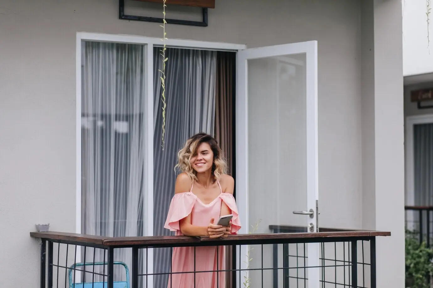 A charming, tanned young woman holding a phone smiles while looking away, dressed in a pink outfit and standing on a hotel balcony.