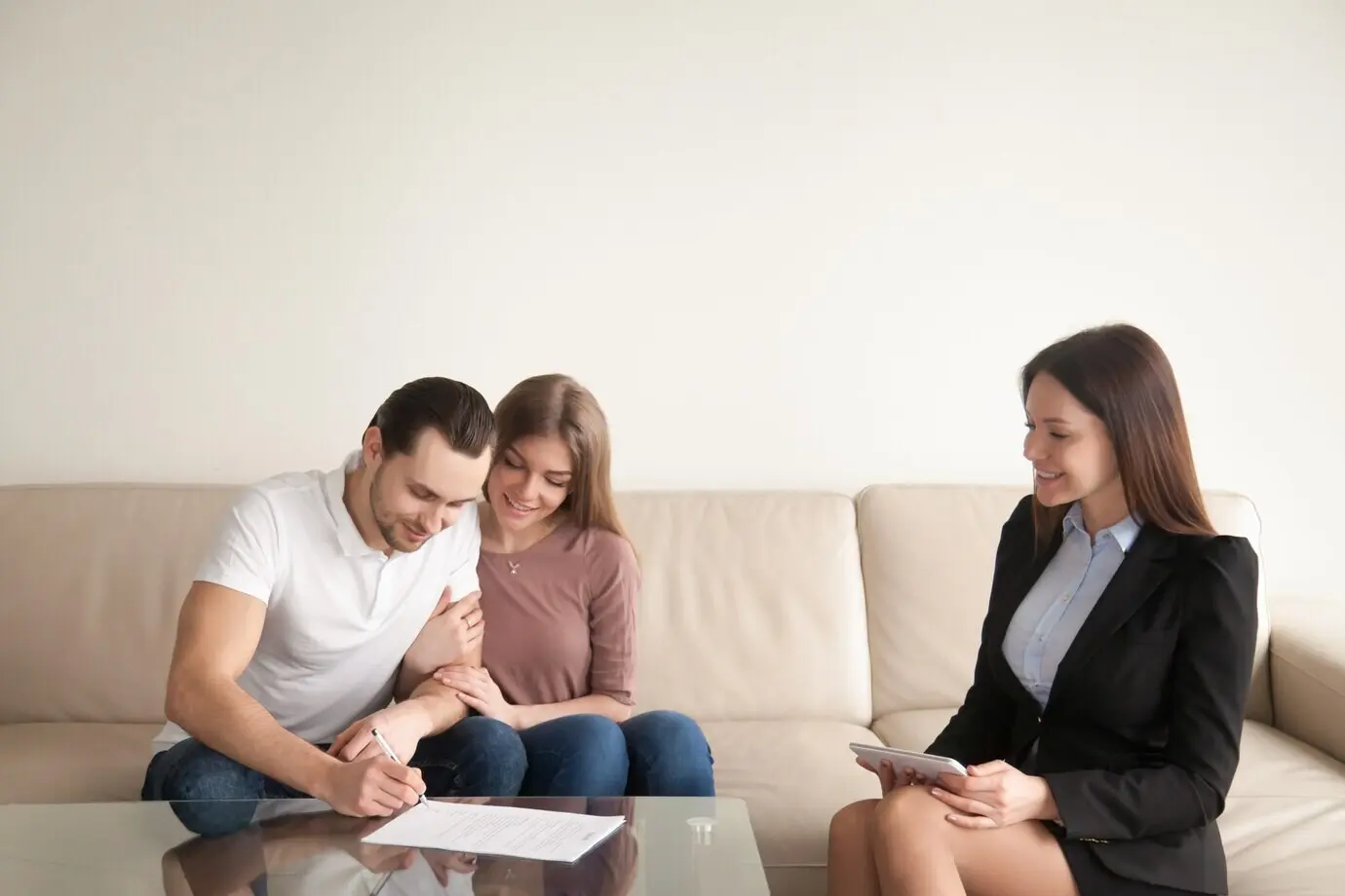 A young man signs papers while seated next to his wife and a realtor.